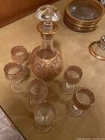 Decanter surrounded by 6 stemmed gold rimmed sherry glasses, and stack of 10 gold rimmed plates.