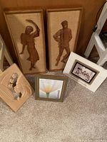 Six mixed media vintage wall art pieces arranged on floor: two carved wooden figures framed in fabric, carved relief plaque, framed floral photo, white photo frame with black and white photo