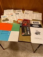 Overview of vintage wargame rulebooks and counters laid out on a table. Several folded rulebooks from different titles visible along with colored paper counters and plastic containers with small game counters.