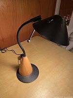 Photo showing angled view of lamp on tabletop, highlighting black metal shade, flexible curved neck, wooden tapered base, and round black metal base plate.