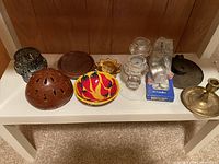Overall view of multiple candle holders and accessories on a white shelf, showing decorative pottery, brass, glass, and metal items.