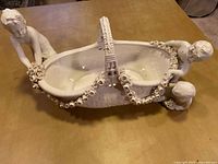 Front angled view showing cherub on the left holding flowers, basket with floral garlands and detailed handle