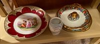 Photo shows four decorative porcelain plates and one small vase on wooden shelf. Plates have classical scenes and fruit motifs with gold trim.