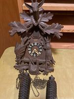 Carved wood cuckoo clock showing front face with Roman numeral dial, stag head at top, rabbit figure on side, bird figures, and pine cone-shaped weights attached to chains.