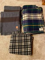 Photo showing folded Hudson's Bay Company grey striped wool blankets, Marshall Field & Company blue green tartan blankets, and the black and white plaid blanket laid on carpet.