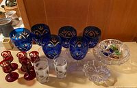 Six blue glasses with cut-to-clear floral patterns, five small red aperitif glasses, two clear etched glasses and two frosted engraved glasses, plus a clear cut glass bowl on the side.