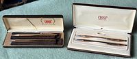 Two vintage Cross pen and pencil sets displayed in their original branded cases on a green surface. The left case is burgundy and gold, the right case is burgundy with cream interior.