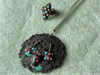 Close-up of pendant necklace and ring showing intricate silver filigree, turquoise and red coral stones arranged in detailed patterns.