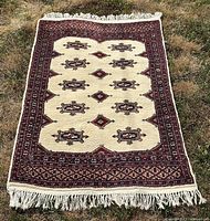 Full view of the vintage handmade Persian rug laid flat on grass, showing entire rug with cream field, red-brown geometric patterned border, and fringed edges.