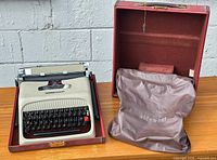 Olivetti Studio 44 typewriter inside maroon carrying case with protective cloth cover beside it, placed on a wooden surface against a gray brick wall.
