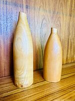Pair of wooden decorative vases or bottles standing on a wooden surface against a wooden background. Taller vase is slender with a narrow top, shorter vase is rounded and squat.