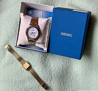 Two Seiko watches shown: men's Solar Quartz watch in blue Seiko box on pillow, and ladies' gold-tone Lassale watch with mesh bracelet laid flat.