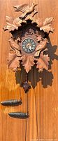 Front view of carved wooden cuckoo clock with birds and leaves, clock face and pine cone weights visible.