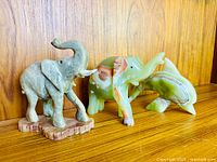 Three carved Mexican onyx animal figurines including two elephants and one dolphin displayed on a wood surface against a wood panel background.