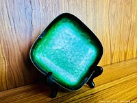 Angle showing the green enamel hammered surface and darker copper edges with patina on the square ceramic dish on display stand.