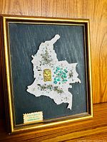 Framed textured map of Colombia with green stones and gold artifact on black background