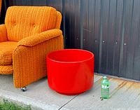 Overall view of glossy red cylindrical planter next to chair and bottle on concrete patio.
