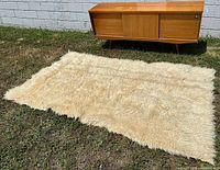 Full view of cream colored long wool fiber Turkish Tülü Kilim rug lying flat on grass next to wooden mid-century sideboard for scale.
