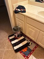 Wide view showing bathroom counter with navy blue Houston Astros towel, two sports themed bath rugs on the floor, and Houston Astros trash can.