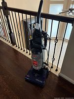 Full view of the upright vacuum cleaner showing dark blue color, upright handle, dust canister, and cord wrapped around handle.