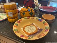 Stack of four pumpkin-shaped orange ceramic bowls, lidded pumpkin-shaped dish with green leaves, large autumn leaf border bowl, two brown ceramic bowls, and assorted plates in background.