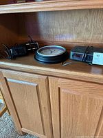 Three clocks on wooden cabinet shelf, including a round wall clock and two alarm clocks with power cords