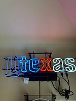 Front view of neon sign spelling 'litexas' illuminated in blue, red, and white colors, mounted on a black metal frame against a wall.