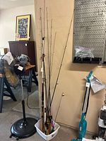 Nine fishing rods grouped together in a white plastic container in a garage. Seven adult rods with cork handles and two shorter children's rods with bright plastic handles.