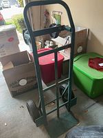 Green painted metal dolly standing in a garage with handle and base plate visible, surrounded by storage bins and boxes.
