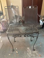 Three piece iron patio set including two mesh metal swivel chairs and one square table with glass top in garage setting with concrete floor.