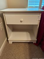 White wooden nightstand close-up showing drawer and open shelf, placed on carpet near wall and red curtain.