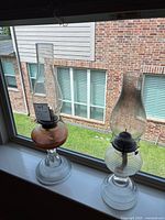 Both vintage oil lamps side by side on a window sill. The amber lamp is slightly shorter than the ribbed clear glass lamp.