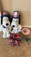 Photo showing two Snoopy patriotic plush toys, one decorative plate partially visible, a patriotic ribbon wreath, and a Texas Longhorn wall decoration on the floor against a beige wall.