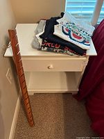 Wooden hat holder rack leaning against a wall beside the nightstand with T-shirts on top.