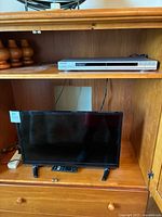 Insignia 23-inch TV on wooden shelf with black remote control in front and silver Sony DVD player on shelf above