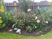 Wide shot of garden bed decor including two turquoise decorative butterfly wall hangings on fence, mosaic sun garden stake, sitting turtle sculpture, cat statue, floral stakes.