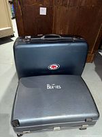 Dark blue American Tourister hard suitcase with red and white circular sticker and black/dark grey Beatles logo suitcase beneath it.