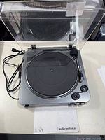 Front view of the Audio-Technica turntable with transparent dust cover open, showing platter, tonearm, and power cord.