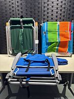 Folded green chaise lounge and two folded beach chairs (blue and striped multi-color) positioned upright side by side showing back carrying straps.