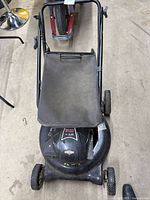 Top-down view of the black gas lawn mower with fabric grass bag attached, showing front wheels and handle bars.