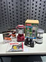 Lot displayed on white table against black woven background showing four camping lanterns, two kayak storage system boxes, binoculars, and packaged poncho.