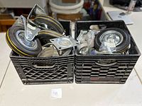 Two black crates containing an assortment of large and small casters, mostly metal with black rubber tires, various mounting types.