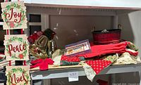 Various Christmas decoration items including a 4-foot square red tablecloth, basket, cardboard signs reading Joy, Peace, Noel, holiday cards, ribbons and fabric items.