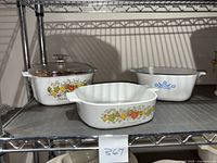Three CorningWare casserole dishes displayed on a wire shelf, two with Spice of Life pattern and one Blue Cornflower