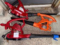 Photo showing three electric leaf blowers together on carpet including red Troy-Bilt, red Toro Ublo blower with vacuum hose, and orange Black & Decker blower