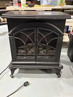 Front view of the black electric heater resembling a wood stove with decorative doors and power cord lying on the floor.