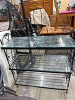 Front angled view showing three glass shelves in a black wrought iron frame with scrollwork on sides and back. Visible chip on glass shelf corner.