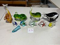Nine assorted glass animal figurines arranged side by side showing variety of animal shapes and colors on a white table.