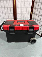Closed rolling toolbox showing its red lid with black handles, two wheels and side handle for rolling.