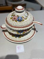 Photo showing large white ceramic soup tureen with hand painted pink and blue flowers, textured floral patterns, and a circular plate underneath. Matching lid and ladle attached.